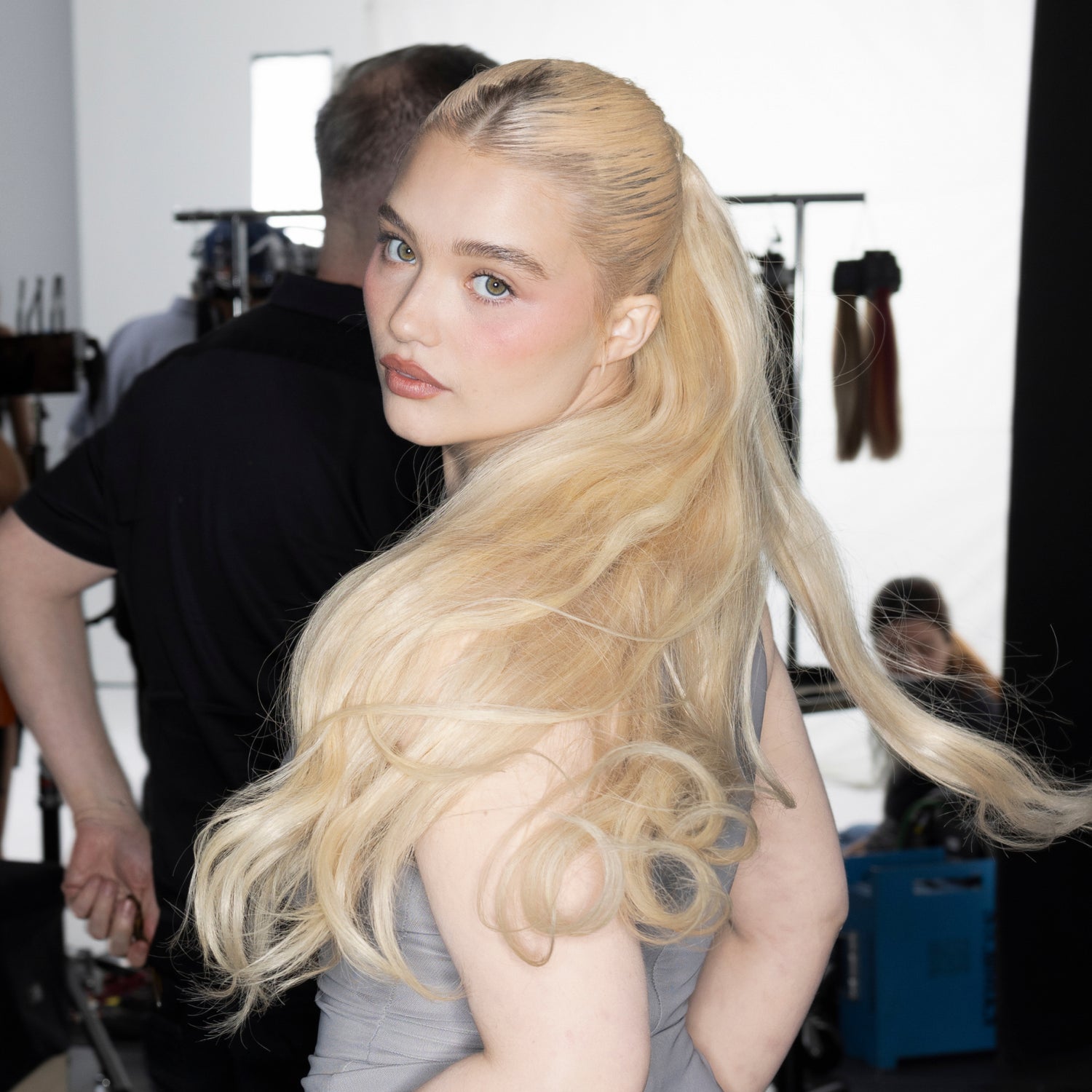 Blonde woman with long, wavy hair in a ponytail, looking over her shoulder in a studio setting.