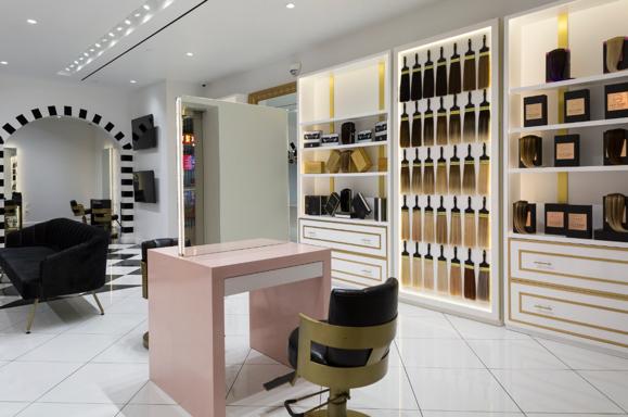 Modern salon with white shelves, a pink styling station, black and gold chairs, white tile flooring.